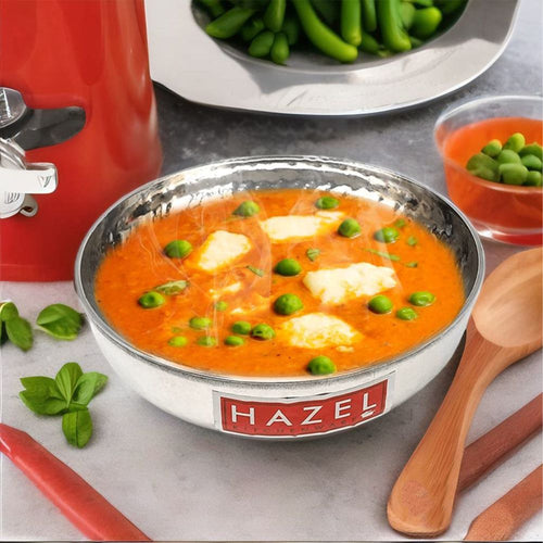 A hammered tasla filled with orange paneer and pea curry, styled on a kitchen counter with a wooden spoon, peas