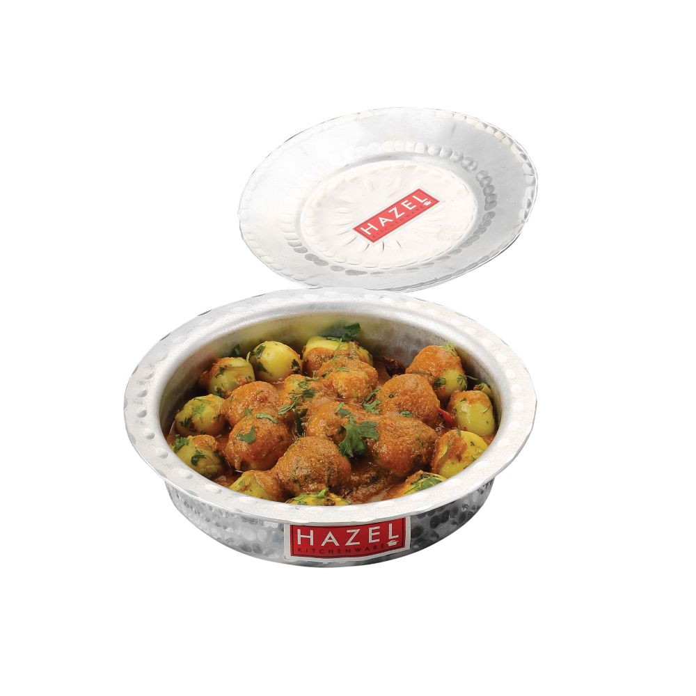 Bhagona filled with potato curry with its lid tilted beside it, shown on a plain white background.