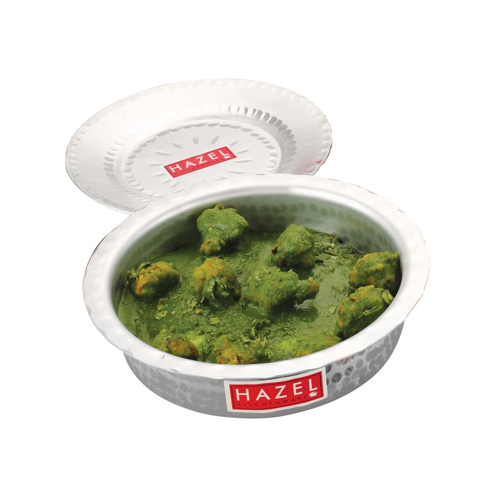 A hammered aluminium dekchi filled with green curry, lid tilted beside it, shown on a white background.