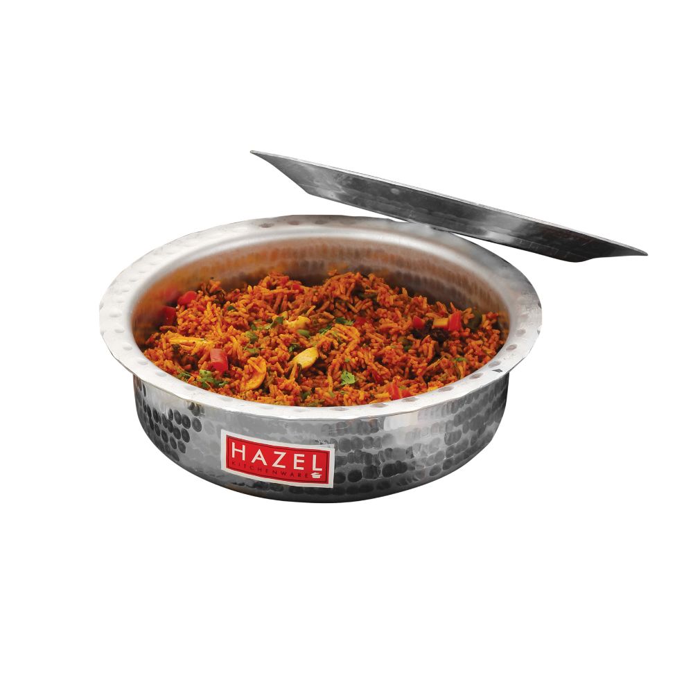 Hammered aluminium patila filled with vegetable curry, lid propped diagonally behind it, on a plain white background.