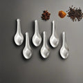 Six small steel masala spoons arranged on a grey surface with scattered whole spices like star anise and peppercorns.