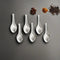 Six small steel masala spoons arranged on a grey surface with scattered whole spices like star anise and peppercorns.
