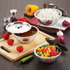 Steel serving bowls holding curry, rice and mixed vegetables on a wooden board surrounded by fresh produce.