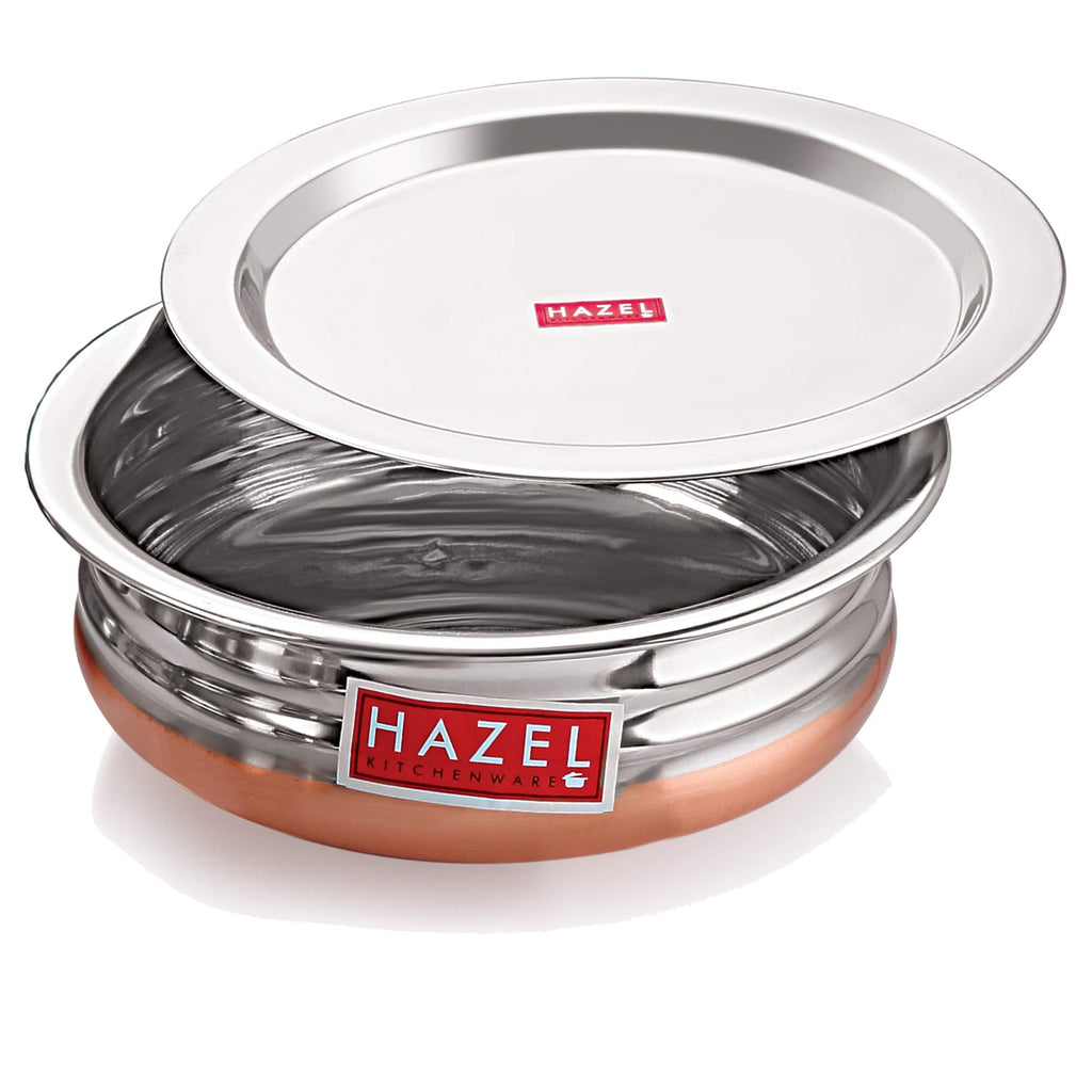 Copper-bottom urli filled with a paneer and pea curry, its lid resting tilted beside it, on a white background.
