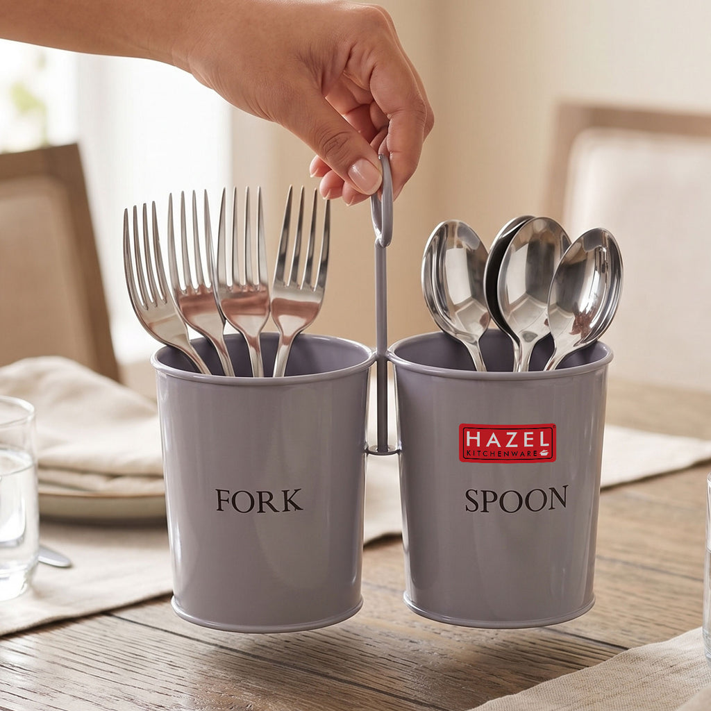 A hand takes a spoon from the grey two-section cutlery holder full of forks and spoons on a wooden table runner.