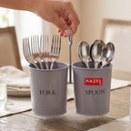 A hand takes a spoon from the grey two-section cutlery holder full of forks and spoons on a wooden table runner.