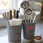 Grey two-section cutlery holder holding knives, ladles, and a spatula on a kitchen counter with a small bowl of sal