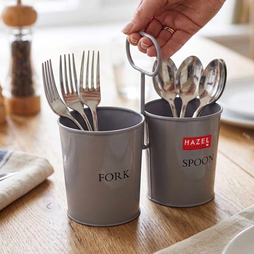 A hand lifts the grey two-section cutlery holder full of forks and spoons off a wooden dining table with a pepper m