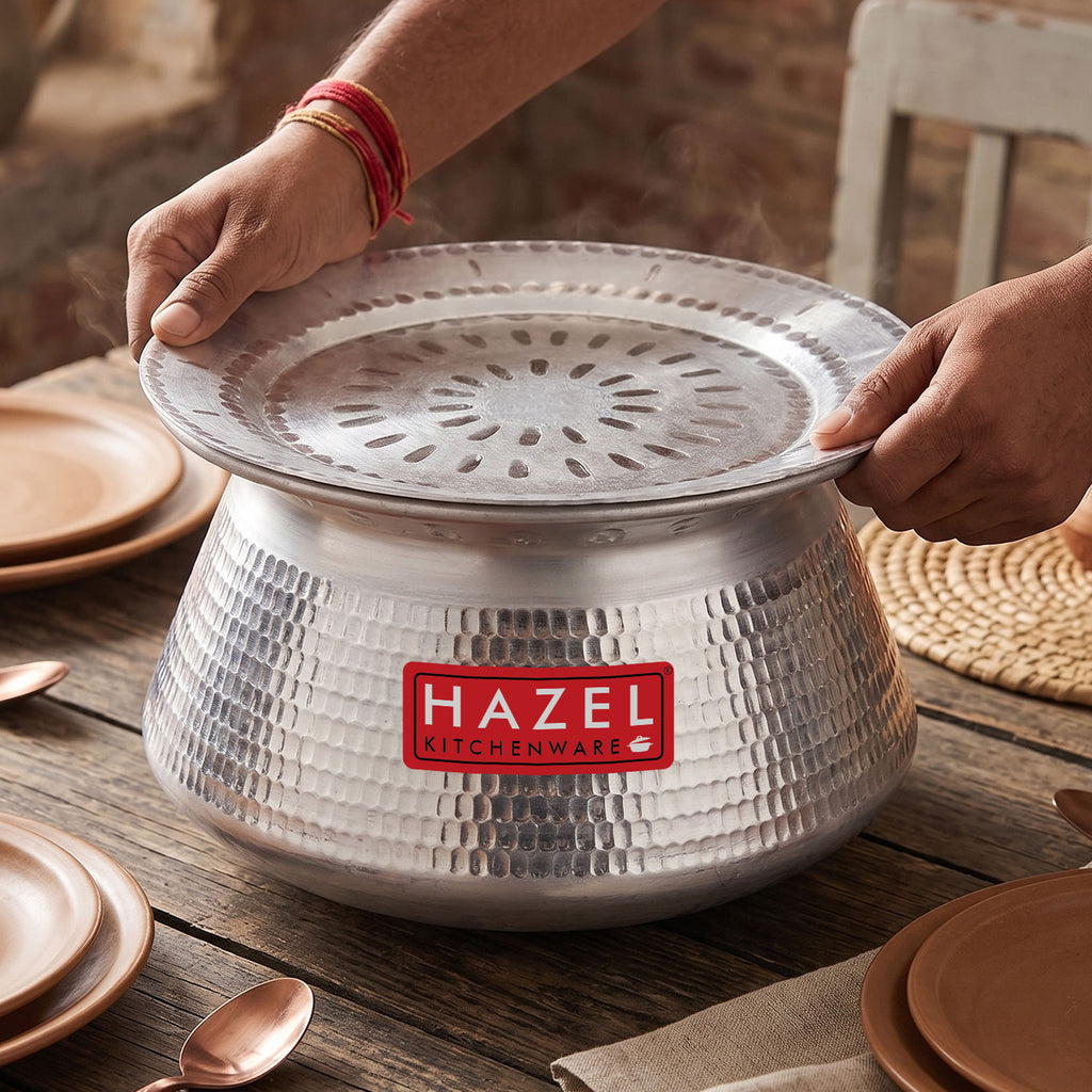 Hands placing a perforated steamer lid onto the aluminium hammered handi, set on a wooden table with plates and cutlery.