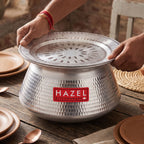 Hands placing a perforated steamer lid onto the aluminium hammered handi, set on a wooden table with plates and cutlery.
