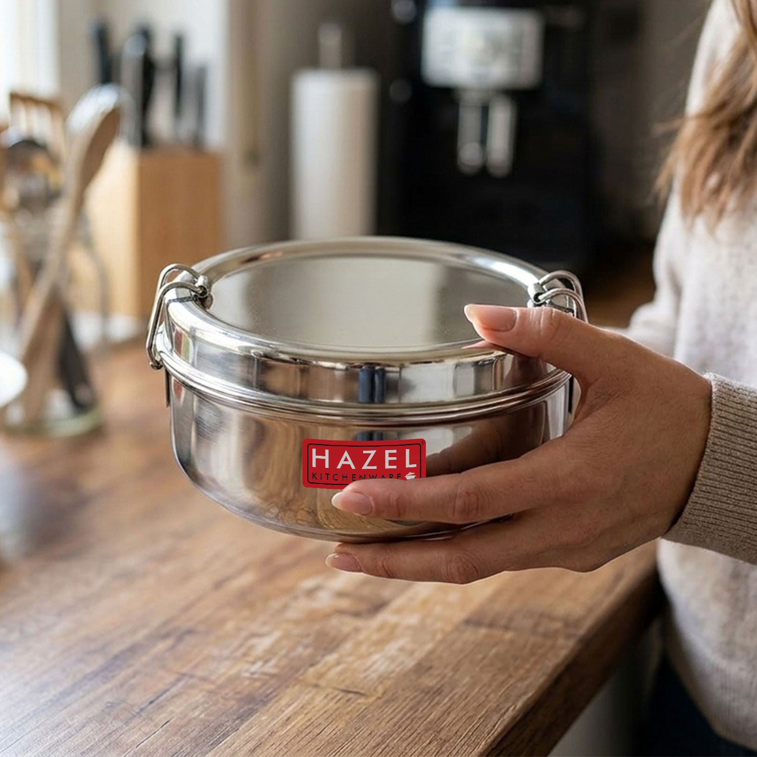 A hand holds a closed stainless steel lunch box in a kitchen setting with a sink and tomatoes visible in the background.